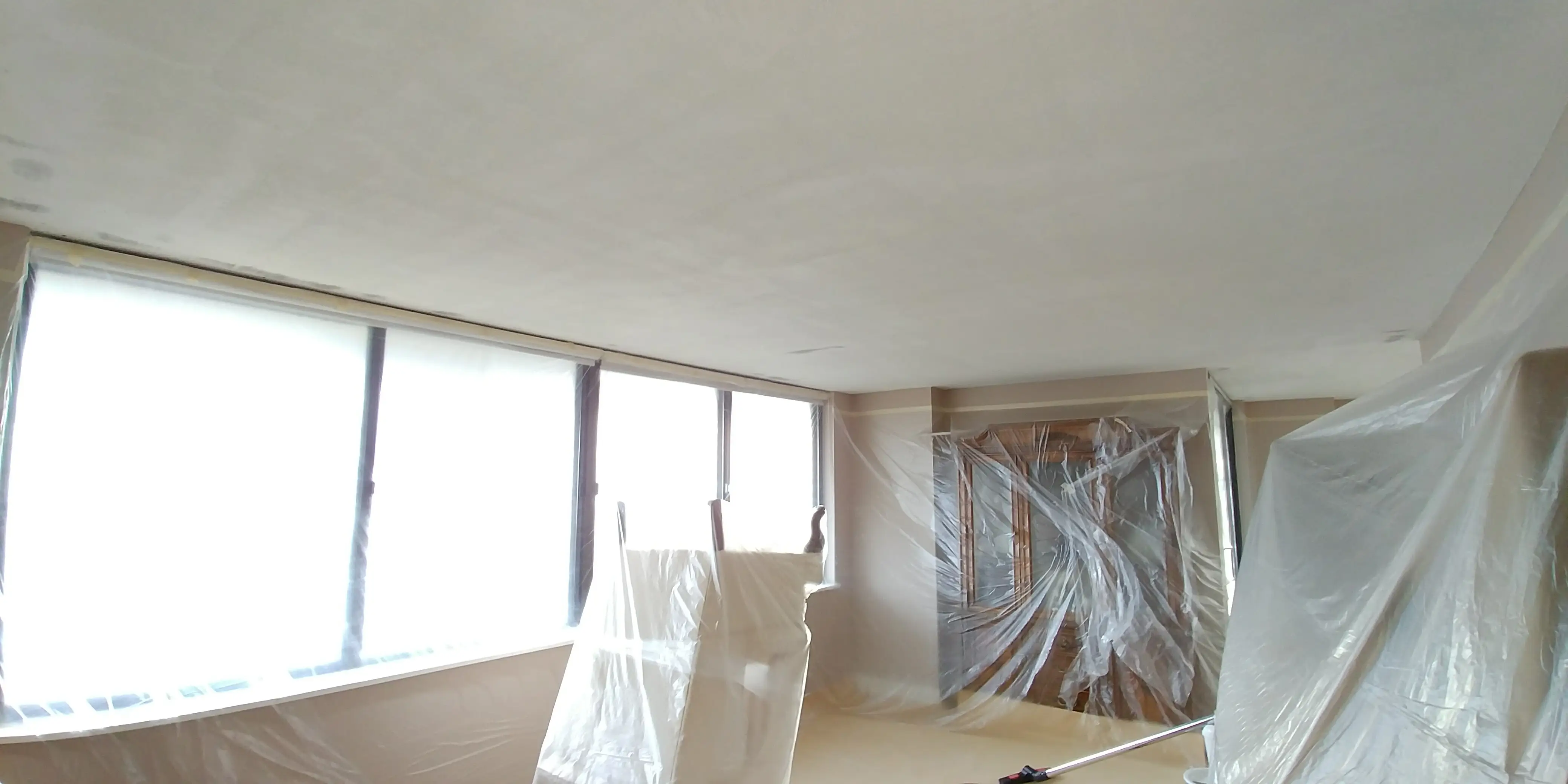 A freshly renovated room with smooth, dust-free ceilings, large windows letting in natural light, and furniture carefully covered for protection during the process.